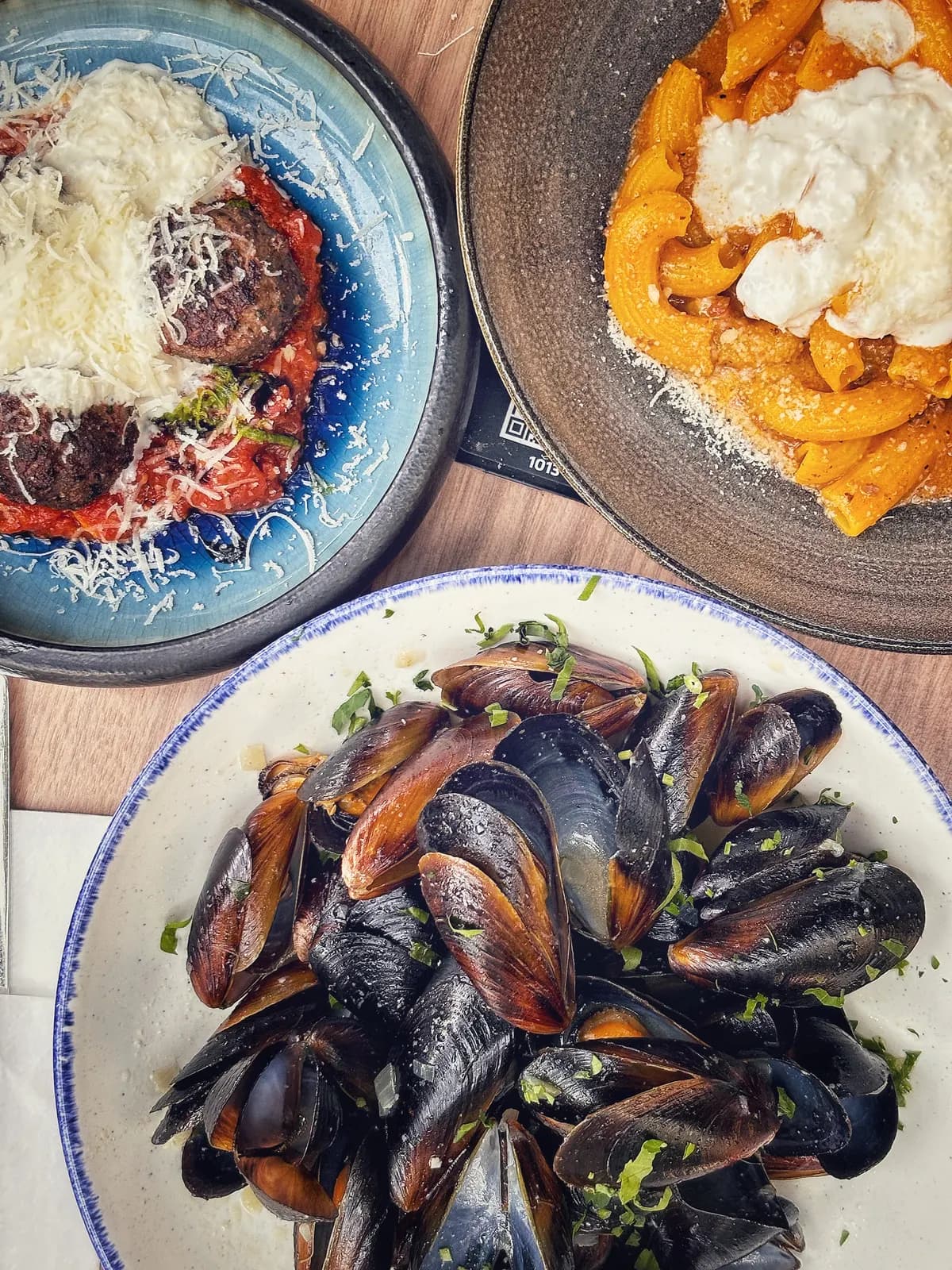 Plate of mussels with herbs beside pasta dishes on a wooden table